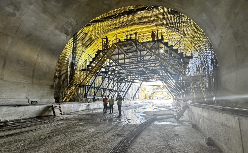 Construction tunnel with steel scaffolding and yellow safety framework; workers wearing hard hats walk inside.