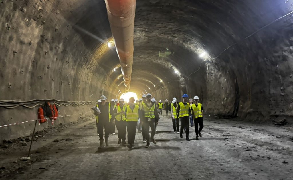 Group of construction workers in hi-vis vests and hard hats walking through a curved underground tunnel with a large pipe overhead and construction lighting at the end