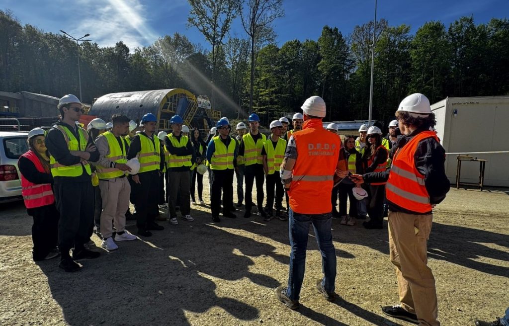 Group of construction workers in high-visibility vests and hard hats listening to two supervisors at an outdoor site briefing into a circle.