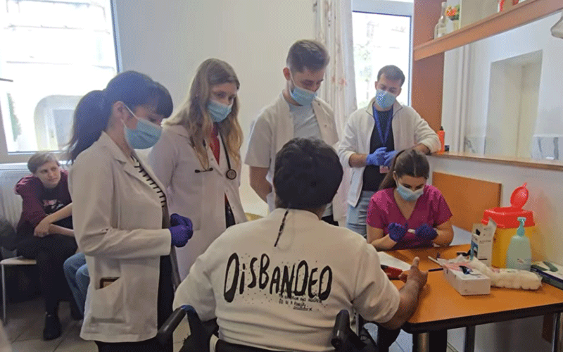 Medical students in lab coats and masks gather around a person in a wheelchair during a clinical training session.