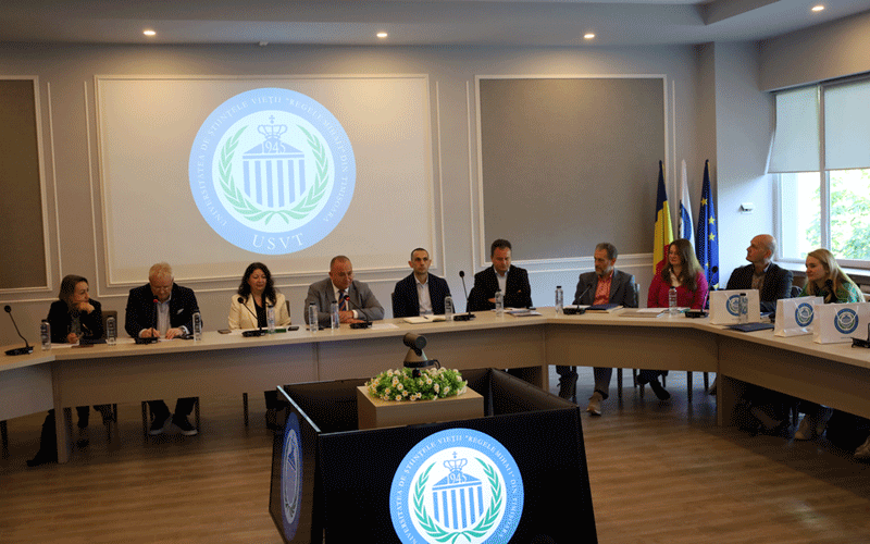 Group of ten people seated around a U-shaped conference table in a formal meeting room, a circular emblem projected on the wall behind them.