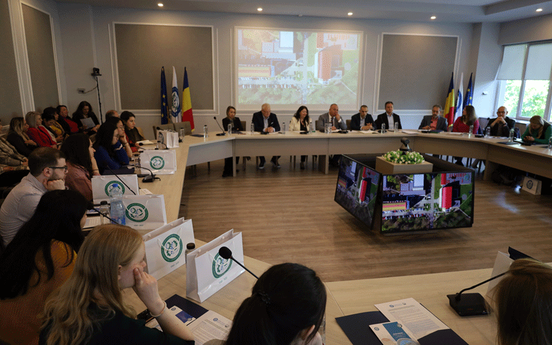 A large conference room with a U-shaped table and about 20 attendees seated around it, flags in the corner, and a projector screen displaying a building image behind the panelists.
