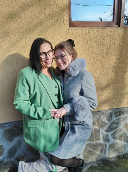 11 - Sursa de Vest Two women pose together outside, smiling; one in a green blazer, the other in a gray coat with fur cuffs, leaning against a wall on a sunny day.