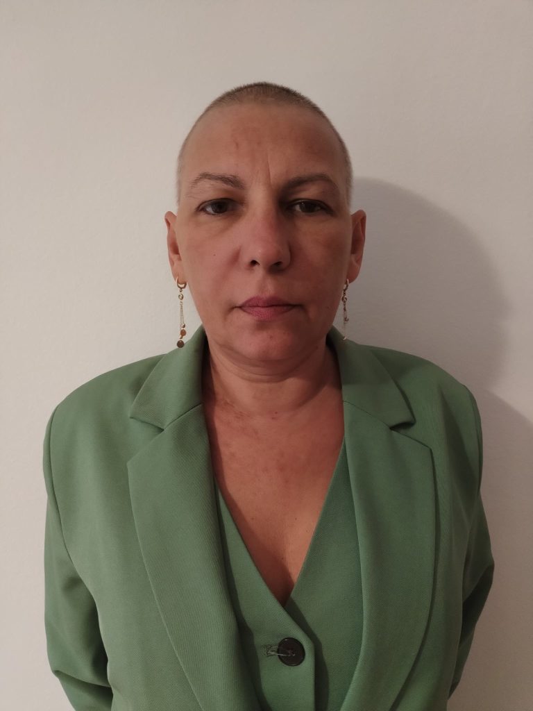 22 - Sursa de Vest Head-and-shoulders portrait of a person with a shaved head, wearing a green blazer and gold earrings against a plain wall, neutral expression.