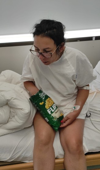 55 - Sursa de Vest Woman in a hospital gown sits on a bed with an IV in her arm, holding a green bag of chips on her lap.
