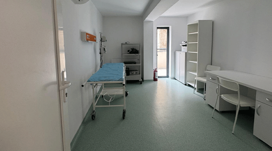 Clinical examination room with a wheeled examination table, medical carts, shelves, and a desk along white walls in a sterile setting.
