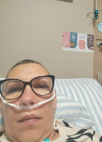 66 - Sursa de Vest Close-up of a person in a hospital bed wearing an oxygen nasal cannula and large black glasses, looking at the camera.