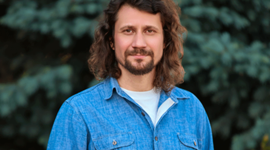 Daniel-Bîcîilă - Sursa de Vest Portrait of a man with shoulder-length brown hair and a short beard wearing a blue denim shirt, standing outdoors with blurred evergreen trees in the background, looking at the camera with a slight smile.