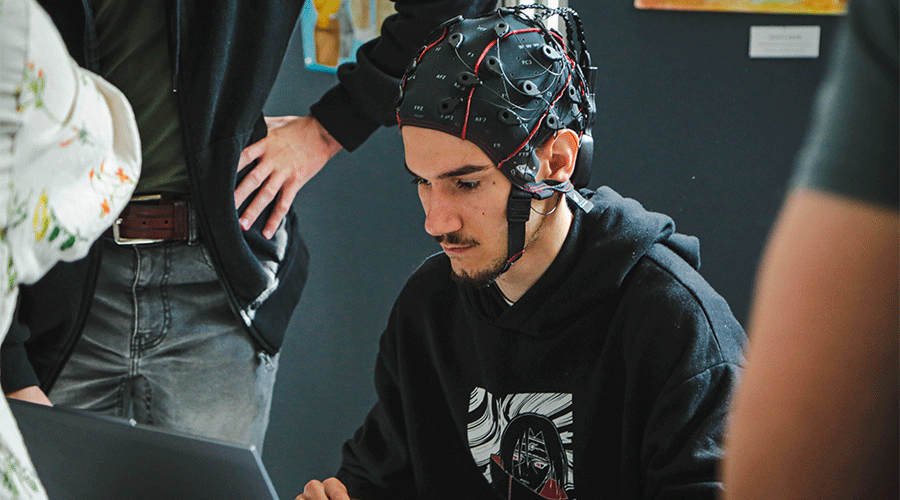 UVT-laboratoare-1 - Sursa de Vest Young man wearing an EEG cap with electrodes, working on a laptop as others observe in a lab setting.