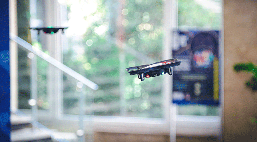 UVT-laboratoare-12 - Sursa de Vest Two small quadcopter drones hovering indoors near glass doors with greenery outside in the background.
