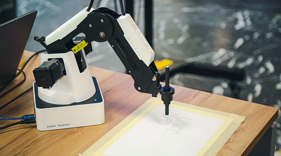 UVT-laboratoare-4 - Sursa de Vest Dobot Magician robotic arm drawing on a sheet of paper taped to a wooden desk with a laptop nearby.