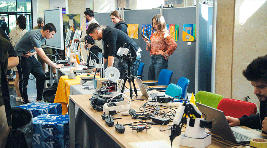 UVT-laboratoare-5 - Sursa de Vest People at a maker-space workshop examining and wiring devices on a long table with laptops and 3D printers in the background