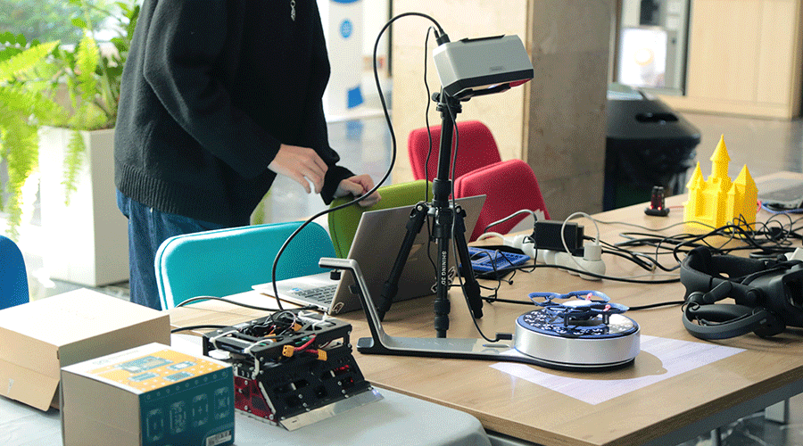 UVT-laboratoare-8 - Sursa de Vest Person standing at a maker table with a tripod-mounted camera, two laptops, cables, and a yellow 3D-printed castle model in the background.