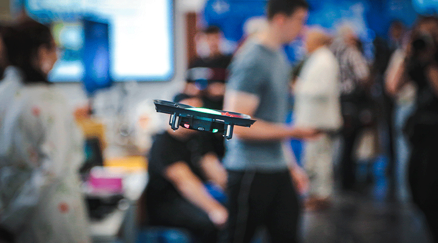 UVT-laboratoare13 - Sursa de Vest A small quadcopter drone hovers indoors at a busy tech expo, with people blurred in the background.