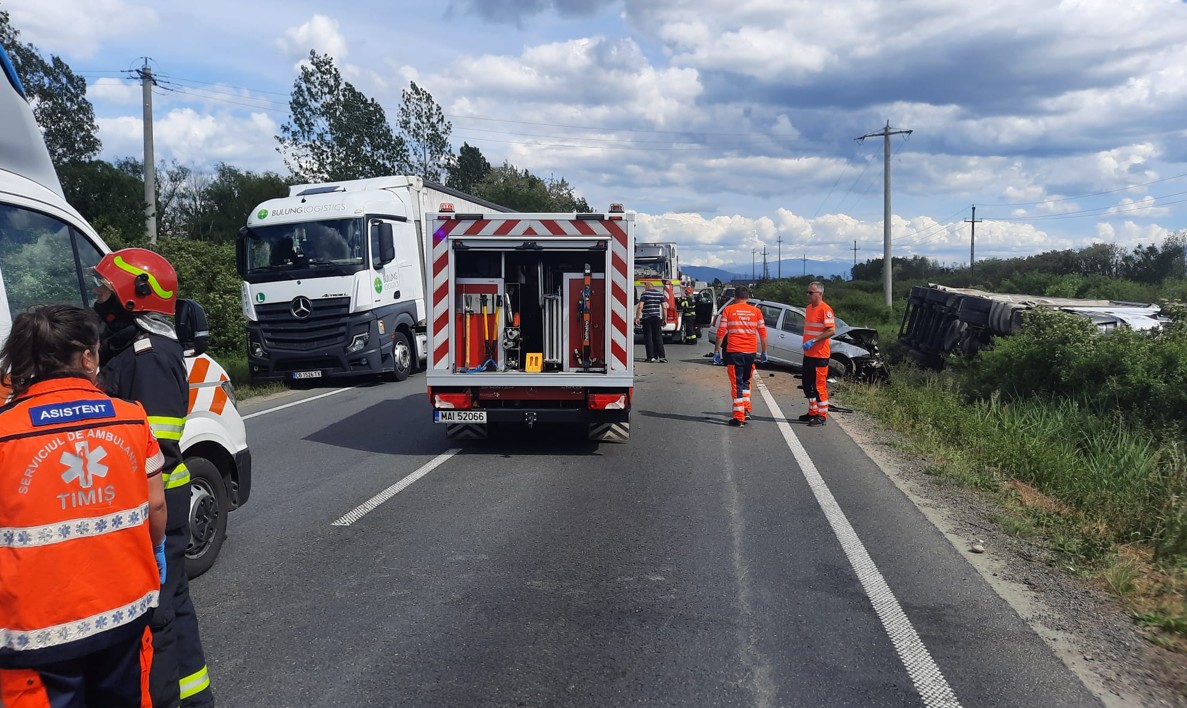 Emergency responders in orange vests at a highway accident scene with a tipped vehicle on the roadside and police/ambulance nearby