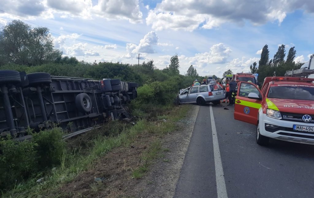 Semi-trailer overturned on the roadside, a silver hatchback blocking the lane, with firefighters and red emergency vehicles at the scene.
