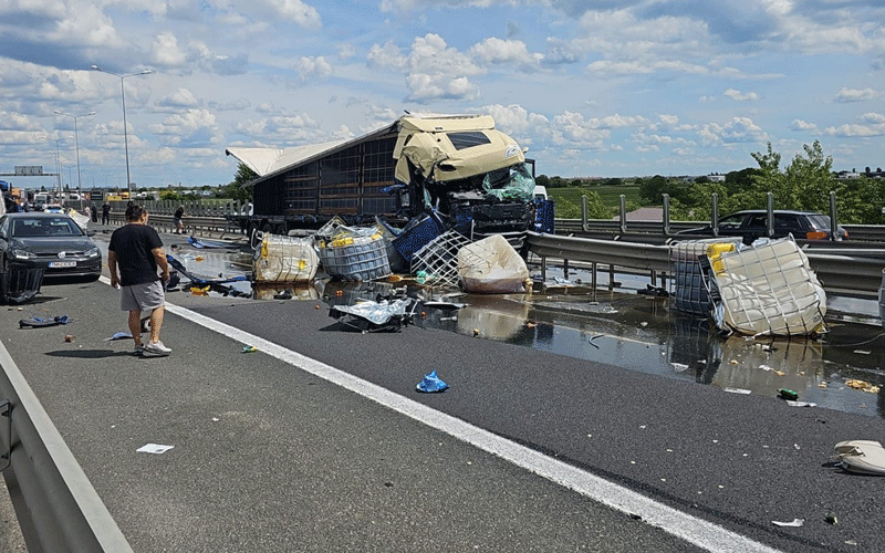 Semi-truck crash on a highway: overturned trailer, scattered pallets and cargo, bystander nearby as cars stop