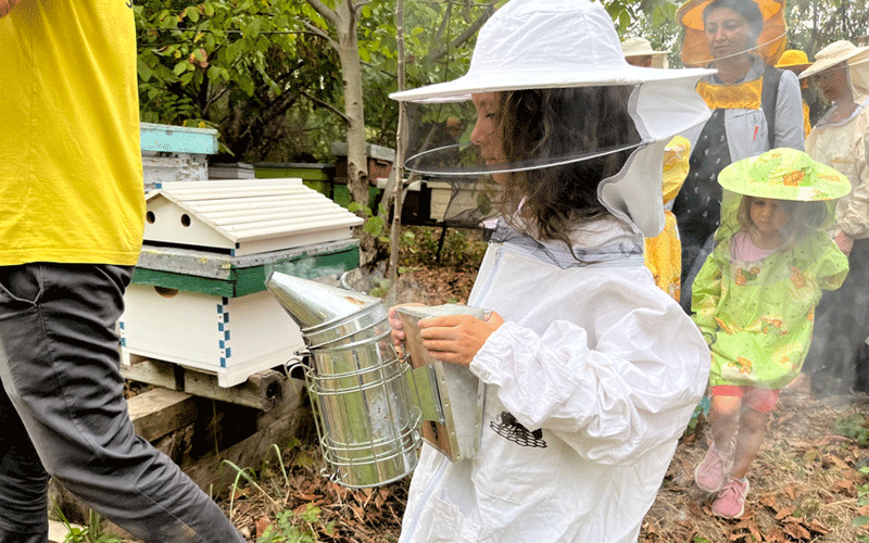 Young girl in a white beekeeper suit holding a smoker at a beekeeping demonstration, with other people and beehives in the background.