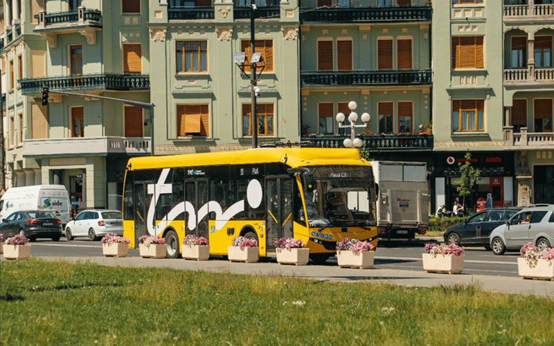Yellow tour bus on a city street in front of pastel green buildings, with flower planters along the curb and parked cars nearby.