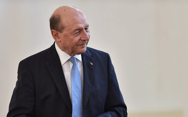 Older man in a dark suit and light blue tie, looking to his left against a neutral backdrop.