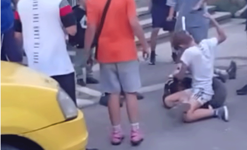 Person kneeling on a city street with arms raised, bystanders nearby and a yellow taxi in the foreground.