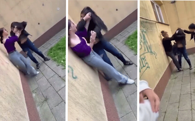 Two young women in casual clothes grapple against a yellow wall outdoors, one in a purple top pulling the other by the arm.