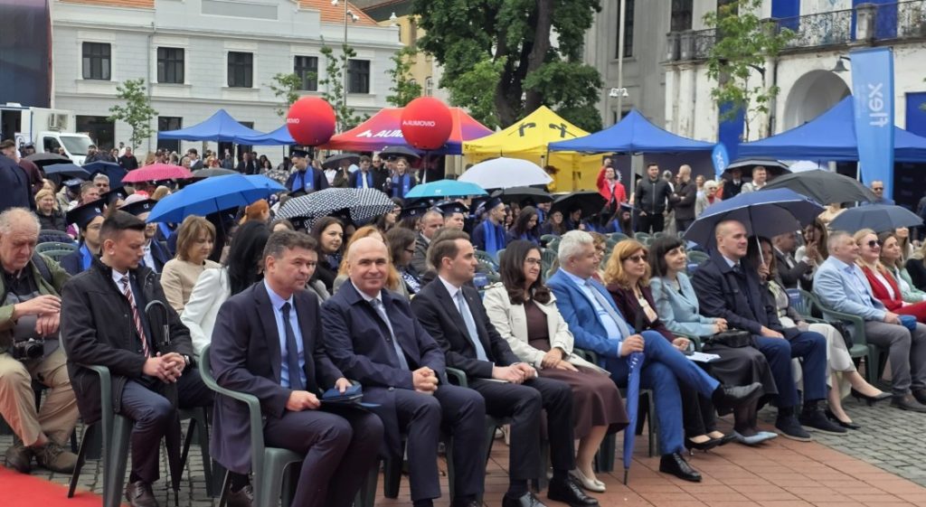 Audience seated at an outdoor event under colorful tents and umbrellas, with several men in suits in the front row and spectators behind them.