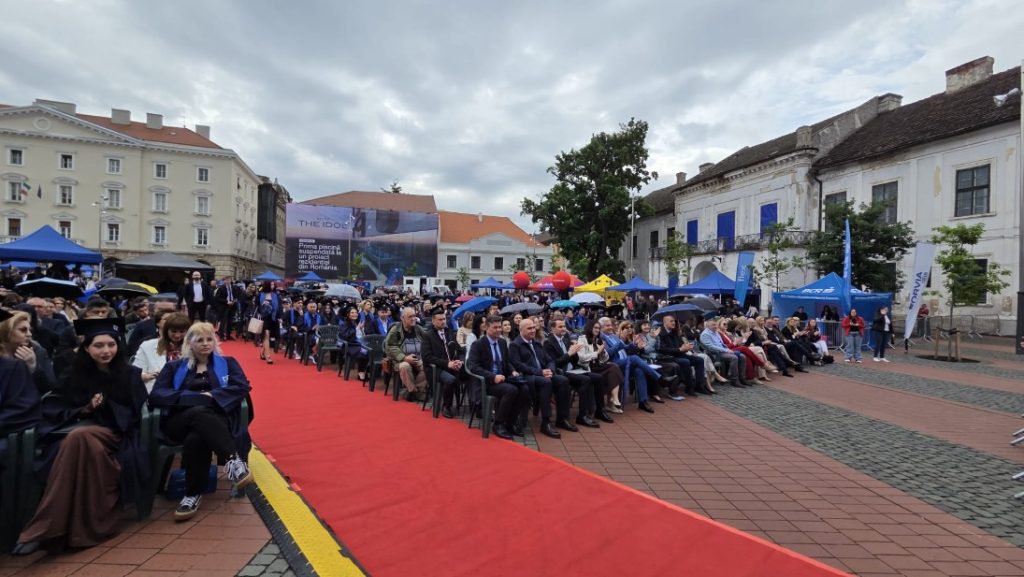 Outdoor ceremony with a red carpet lined by seated attendees in city square, tents and banners in the background under a cloudy sky.