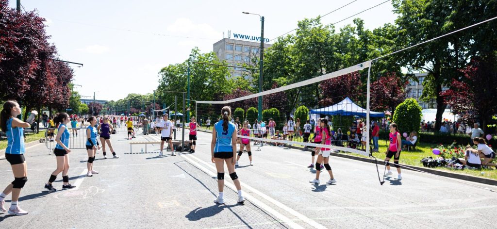 bulev12-1536x708 - Sursa de Vest Outdoor street volleyball game with players in blue and pink uniforms, net set up on a sunny road and spectators on the sidelines.