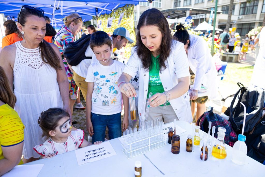 bulev13-1536x1024 - Sursa de Vest A woman in a white lab coat demonstrates science experiments at an outdoor booth for children, with test tubes and bottles on the table and curious onlookers nearby.