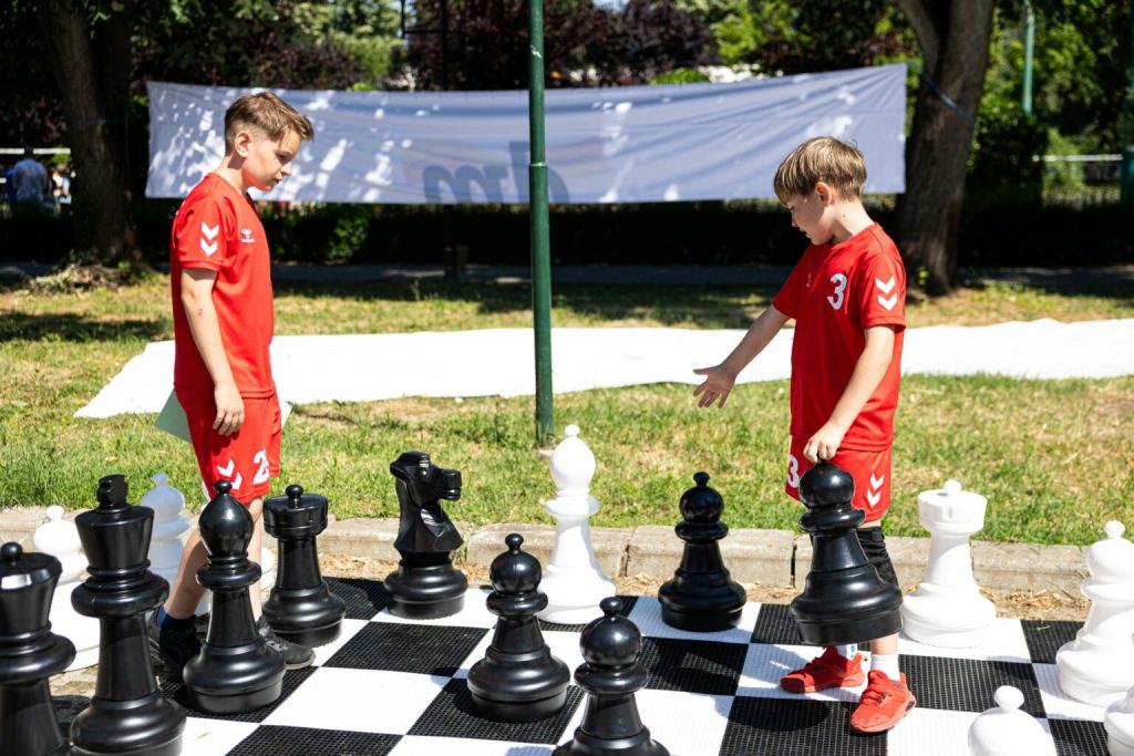 bulev16-1536x1024 - Sursa de Vest Two boys in red sports uniforms play giant outdoor chess on a black-and-white board in a park, facing each other with large pieces between them, one extending a hand toward a piece.
