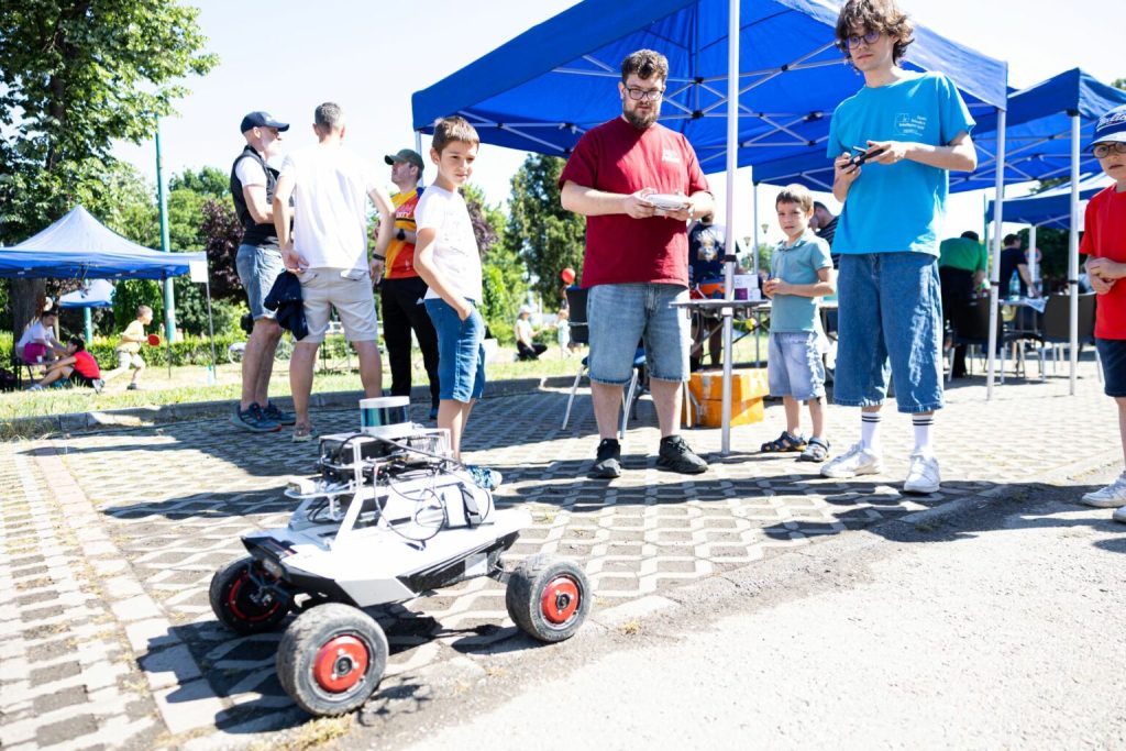 bulev21-1536x1024 - Sursa de Vest A white remote-controlled car on a paved surface with exposed wiring, surrounded by kids and adults at an outdoor event under blue tents.