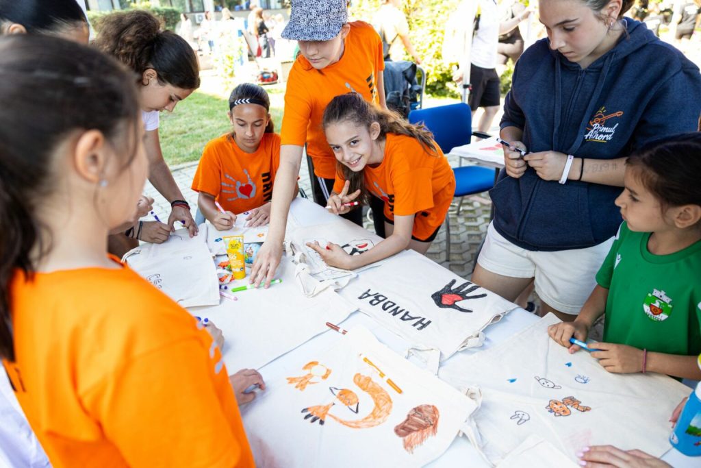 bulev30-1536x1024 - Sursa de Vest Group of kids and teens painting designs on white tote bags at an outdoor event; several wear orange shirts in a sunlit park.