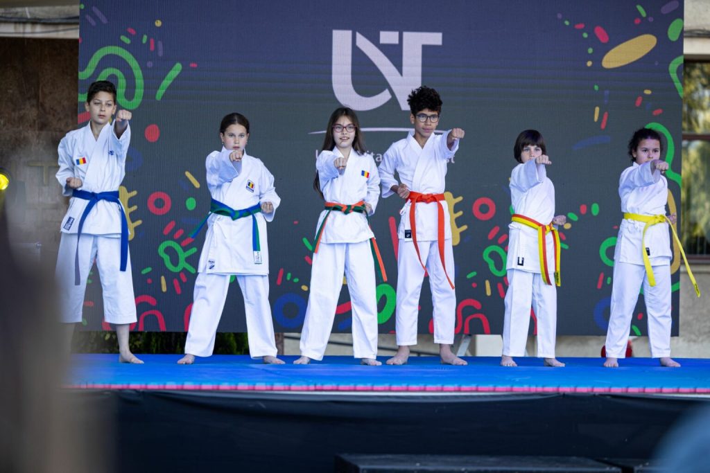 bulev34-1536x1024 - Sursa de Vest Seven children in white karate uniforms with colored belts stand in a line on a blue stage, delivering front punches in a coordinated display.