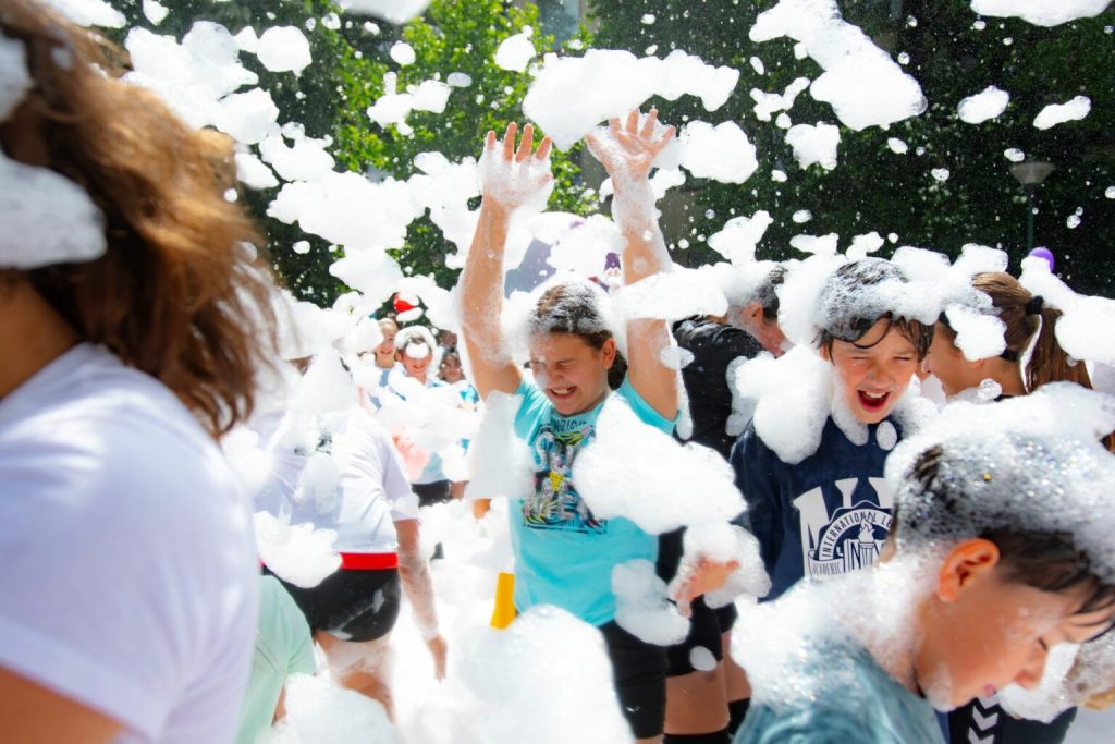 bulev37-1536x1024 - Sursa de Vest Children play in a foam party outdoors, hands raised as soap bubbles swirl around them, smiles on their faces.