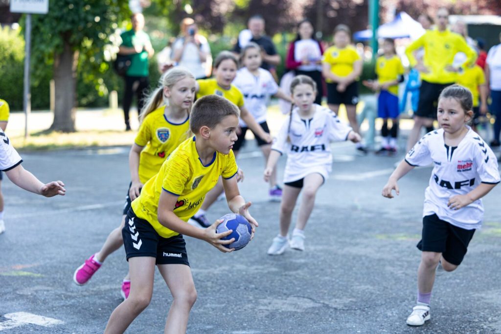 bulev9-1536x1024 - Sursa de Vest Young kids in yellow and white jerseys playing handball on an outdoor court, a boy in yellow holds a blue ball and runs forward while others chase him.