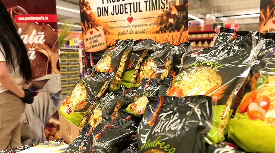Pile of snack bags in a supermarket display, black and green packaging stacked on a table, with a blurred person in white on the left and store signage in Romanian in the background.