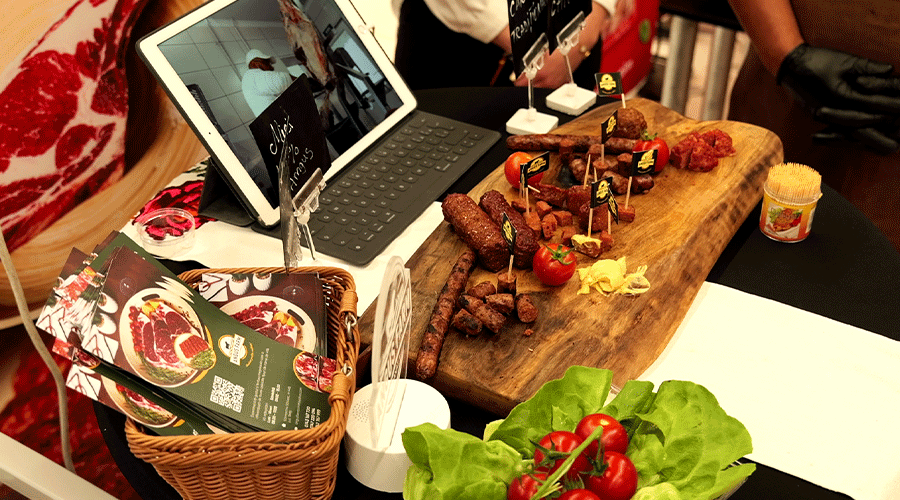Assorted grilled meat skewers and small bites on a wooden board at a tasting booth, with a tablet display in the background.