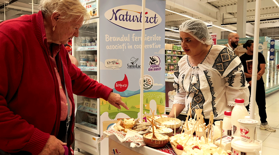 Older man in a red jacket points at a vendor table full of bread, pastries, and skewers as a woman in traditional embroidered clothing stands behind it in a store.