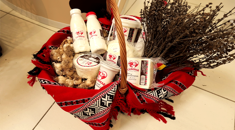 Gift basket on a tiled floor, wrapped in red patterned cloth, filled with white bottles, cups, garlic bulbs, and dried stems/branches inside the basket.