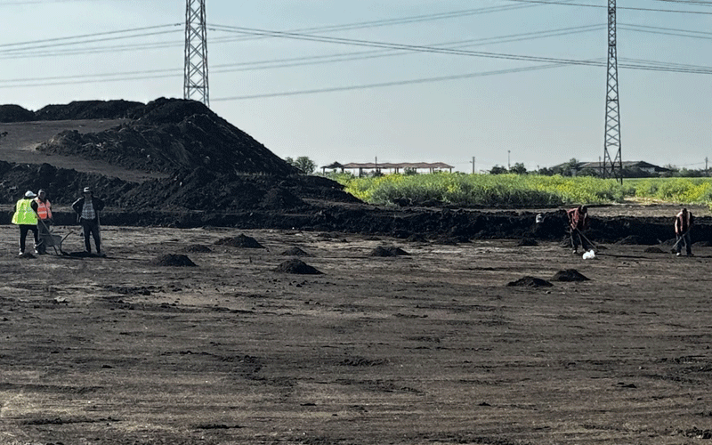 Construction site with workers spreading dark soil piles on a barren field, power lines overhead.