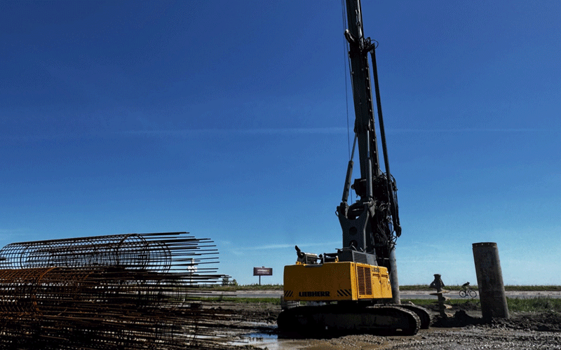centura-vest - Sursa de Vest Yellow Liebherr drilling crane on a muddy construction site with stacked rebar to the left and a clear blue sky above.