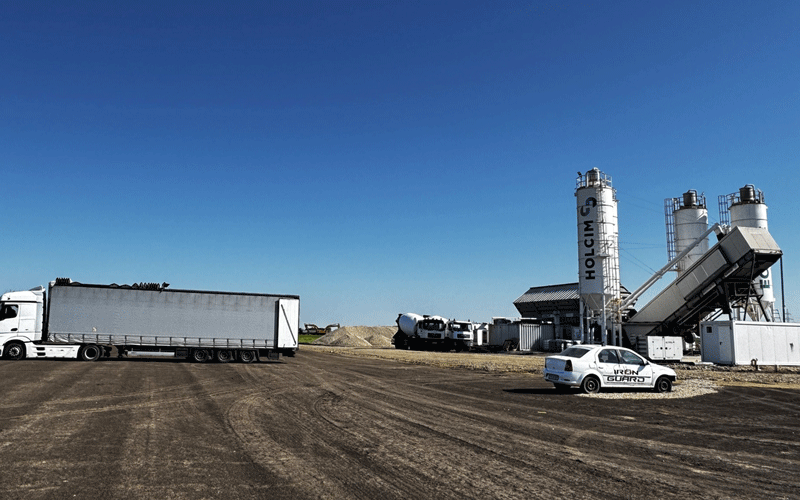 centura-vest4 - Sursa de Vest Industrial plant with tall silos and a diagonal conveyor, white trucks and a small car on a dirt lot under a clear blue sky (Holcim facility)