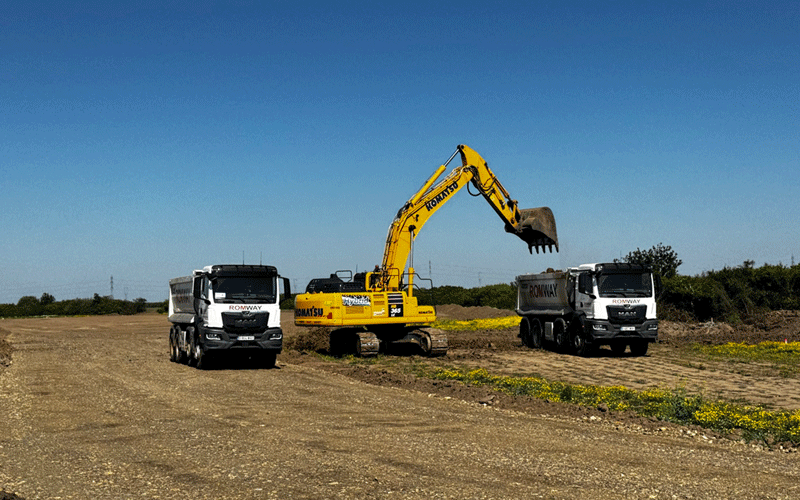 centura-vest5 - Sursa de Vest Yellow excavator loads dirt into a white dump truck while another dump truck waits on a dusty construction site under a clear blue sky.