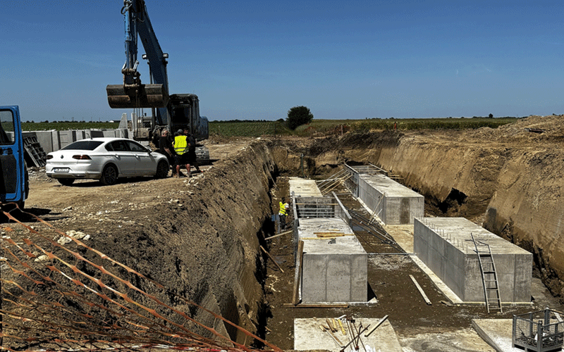 Construction site with a large excavator and a white car near the edge of a deep trench; workers in high-visibility vests.