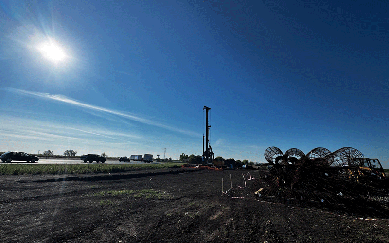 centura-vest6 - Sursa de Vest Construction site along a highway with a tall drilling rig and coiled metal pipes on the right; bright sun in a blue sky.