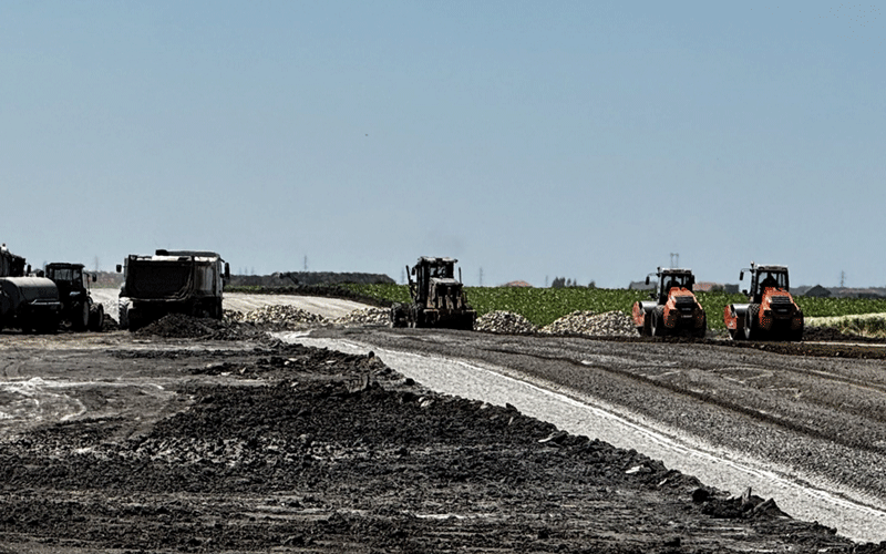 centura-vest9 - Sursa de Vest Road construction scene with several orange compactors and a paving machine laying asphalt along a dirt-covered roadway; piles of gravel on the sides under a clear sky.
