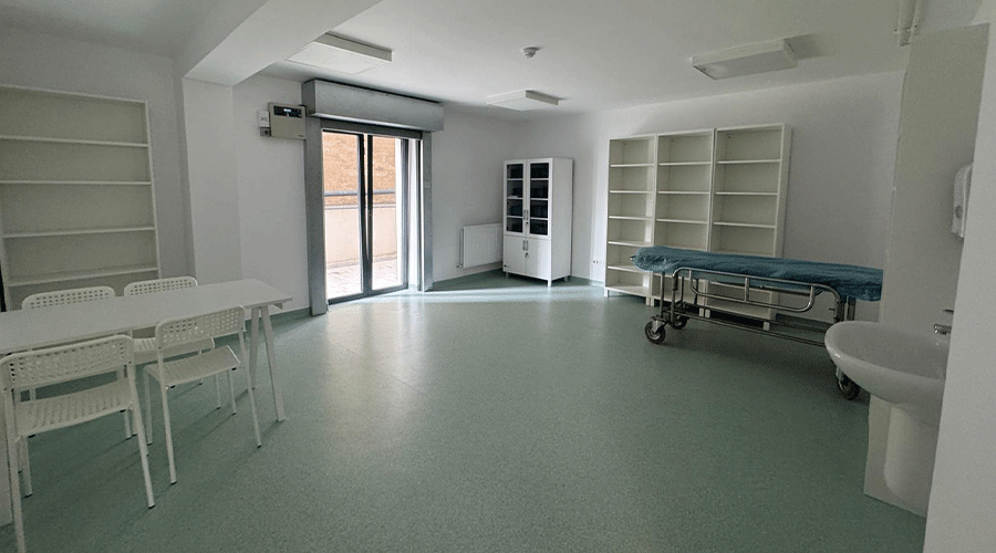 Bright, empty clinic room with white shelves, a wheeled exam bed, and a glass door leading outside on a pale green floor.