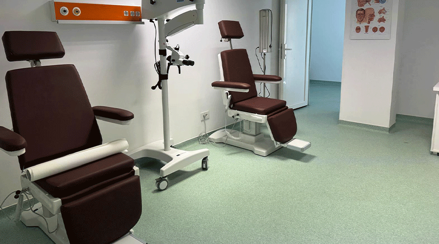 Two burgundy medical examination chairs in a clean clinic room with a green floor and nearby equipment stand.
