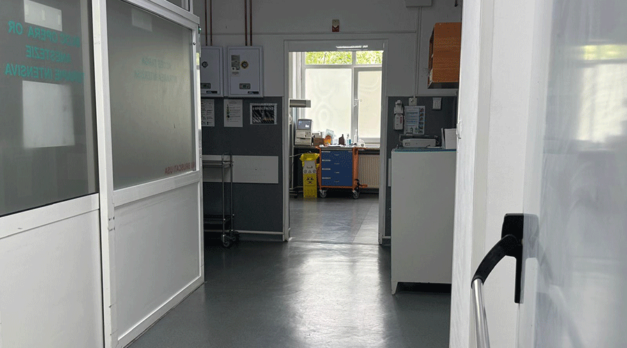 Hospital corridor viewed from an open doorway, leading to a room with medical equipment and a yellow biohazard bin in the back area.